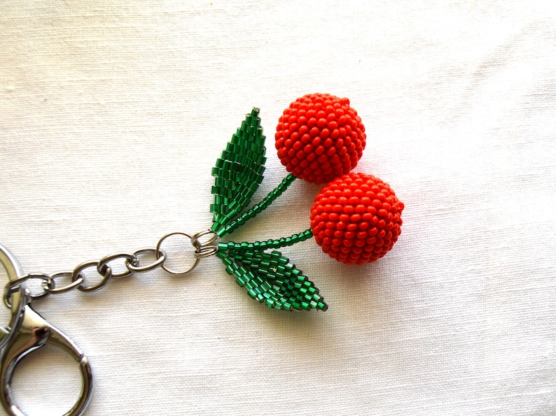 May include: A beaded cherry keychain with a silver chain and green leaves. The cherries are red and made of small beads.