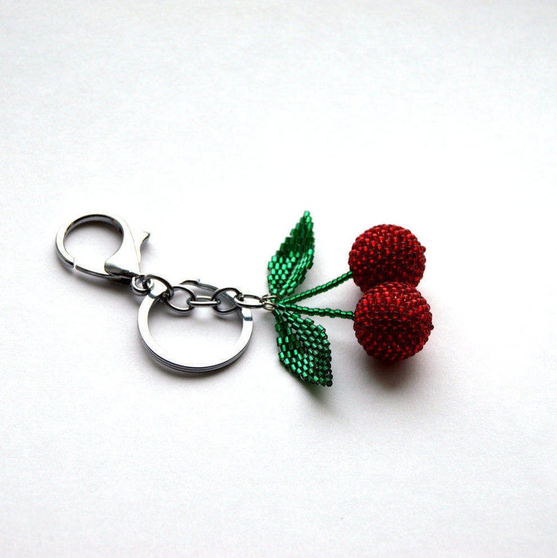 May include: A silver key chain with a beaded cherry charm. The cherry is red and has green leaves.