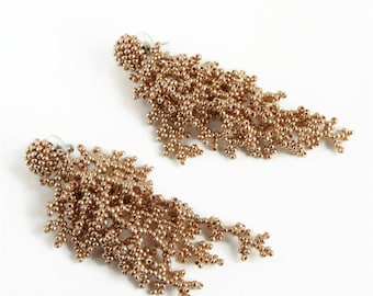 May include: A pair of gold-coloured beaded earrings. Each earring has a cluster of small beads at the top, with cascading strands of beads forming a fringed design. The earrings are set against a white background.