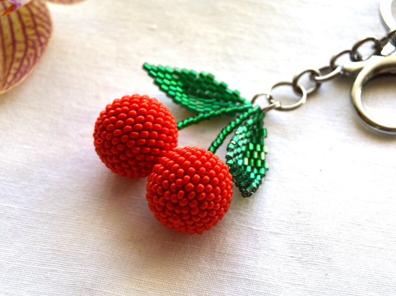 May include: A beaded cherry keychain with two red cherries and green leaves. The keychain is attached to a silver metal ring.