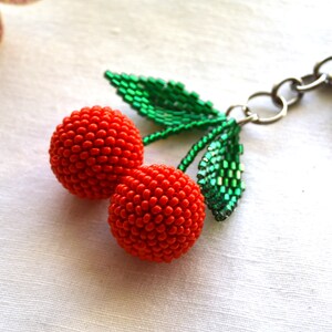 May include: A beaded cherry keychain with green leaves and a silver metal ring. The cherries are red and made of small beads.
