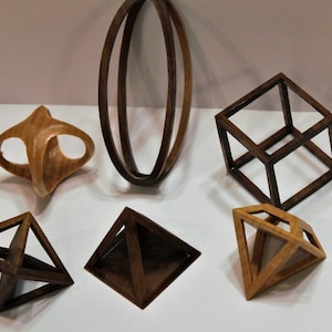 May include: Six wooden geometric shapes, including a cube, a pyramid, and a sphere. The shapes are made of different types of wood and have a natural finish.