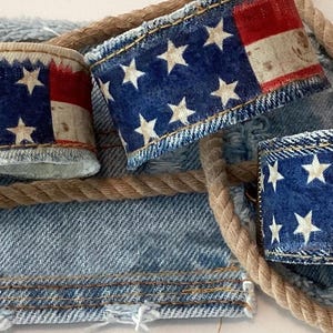 May include: Three wide cuff bracelets featuring a printed American flag design with white stars on a blue field and red and white stripes. The cuffs have visible stitched edges and a textured appearance, arranged alongside a length of natural tan rope.