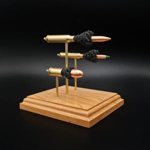 May include: A display stand featuring three horizontal bullet-shaped objects mounted on brass rods attached to a square wooden base. Each bullet casing is gold-coloured with a copper-toned tip, accented by a black textured knot detail.