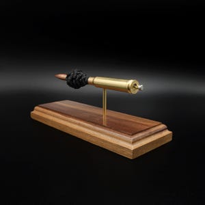 Puede incluir: Una pieza decorativa que presenta un casquillo de bala de color cobre con un acento de cord&oacute;n negro anudado, montado horizontalmente sobre una fina varilla de lat&oacute;n. El conjunto est&aacute; fijado a una base rectangular de madera pulida.