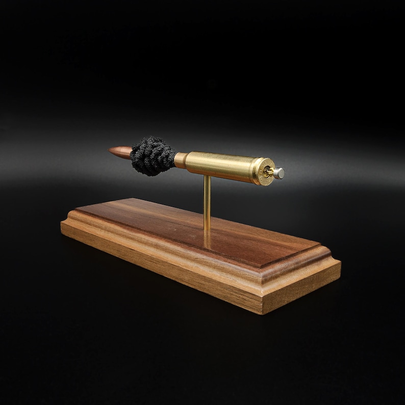 Puede incluir: Una pieza decorativa que presenta un casquillo de bala de color cobre con un acento de cord&oacute;n negro anudado, montado horizontalmente sobre una fina varilla de lat&oacute;n. El conjunto est&aacute; fijado a una base rectangular de madera pulida.