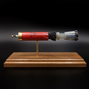 May include: A red shotgun shell casing mounted on a gold-coloured metal rod and a rectangular wooden base. The shell features a brass base, a black cord wrapped around the centre, and a clear plastic wad containing small metallic pellets at the end.