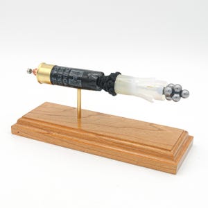 May include: A black and gold shotgun shell with a white plastic casing and a cluster of silver balls at the end. The shell is mounted on a wooden stand.