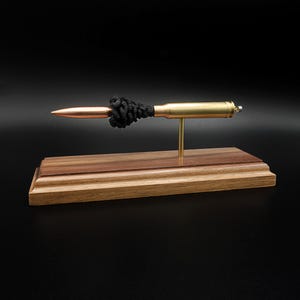 Puede incluir: Una pieza decorativa que presenta un cartucho de bala met&aacute;lico con punta de color cobre y un detalle de nudo negro texturizado, montado horizontalmente sobre una fina varilla de lat&oacute;n sujeta a una base rectangular de madera pulida.