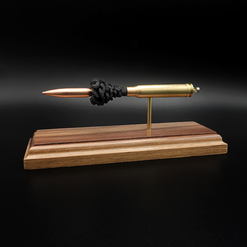 Puede incluir: Una pieza decorativa que presenta un cartucho de bala met&aacute;lico con punta de color cobre y un detalle de nudo negro texturizado, montado horizontalmente sobre una fina varilla de lat&oacute;n sujeta a una base rectangular de madera pulida.