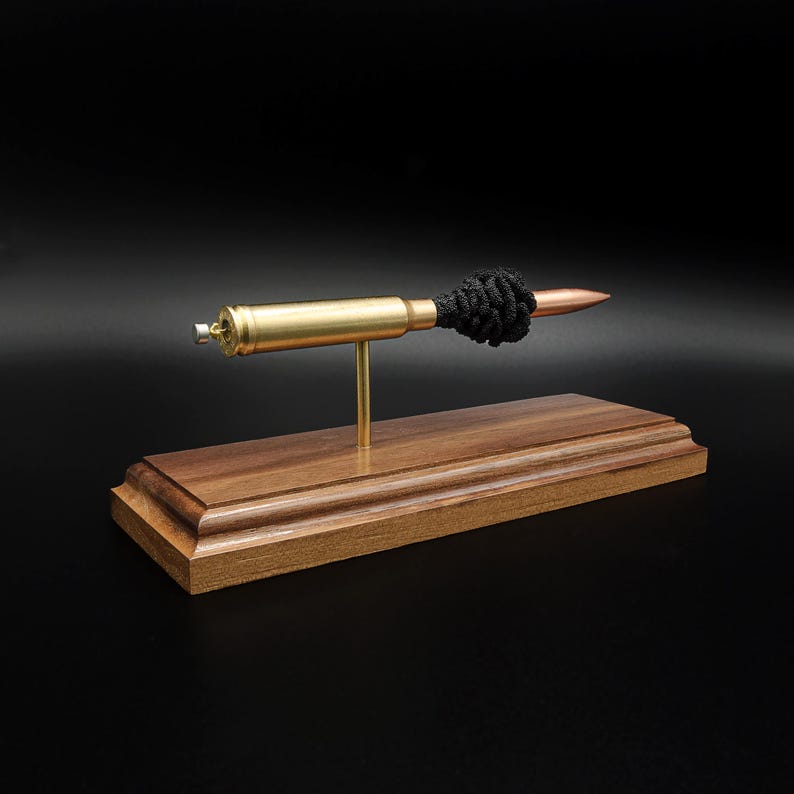 Puede incluir: Un bol&iacute;grafo hecho con un casquillo de bala de lat&oacute;n con punta de cobre y un adorno de cuerda negra anudada, montado horizontalmente sobre una varilla de lat&oacute;n sujeta a una base de madera rectangular con bordes biselados.