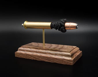 Può includere: Un oggetto decorativo composto da un bossolo in ottone e una punta di proiettile color rame collegati da un materiale nero testurizzato, montato su un sottile stelo in ottone fissato a una base rettangolare in legno con venature.