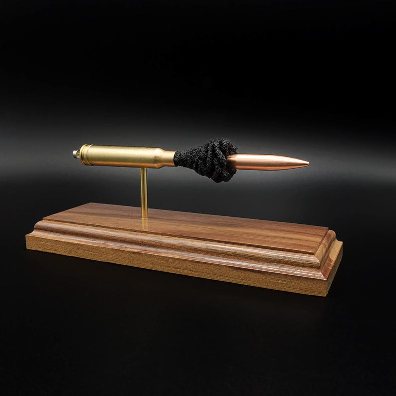 Puede incluir: Un expositor decorativo con un objeto horizontal en forma de bala con casquillo color lat&oacute;n y punta color cobre, separados por un cord&oacute;n negro anudado. El objeto est&aacute; montado sobre un poste fino de lat&oacute;n en una base de madera.