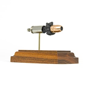 May include: A silver and copper bullet with a black powder charge is displayed on a wooden stand. The bullet is mounted on a metal rod that is attached to the stand. The stand is made of dark wood with a smooth finish.