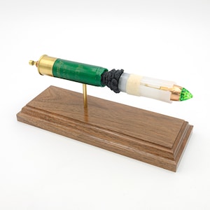 Pode incluir: Uma peça de arte feita a partir de uma bala de espingarda desmontada. A bala é verde e dourada, com uma secção preta e uma ponta transparente, branca e cor de cobre. É montada numa base de madeira com uma haste de latão.