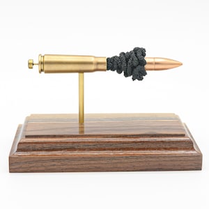 May include: A decorative sculpture featuring a brass bullet with a copper tip, adorned with black beads. The bullet is mounted on a gold-colored stand and set on a wooden base, showcasing a unique blend of materials and textures.