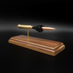 Puede incluir: Un expositor decorativo que presenta un casquillo de bala horizontal de lat&oacute;n y cobre montado sobre una fina varilla de metal dorado. La bala est&aacute; acentuada con un detalle de nudo negro texturizado y descansa sobre una base rectangular de madera marr&oacute;n pulida.