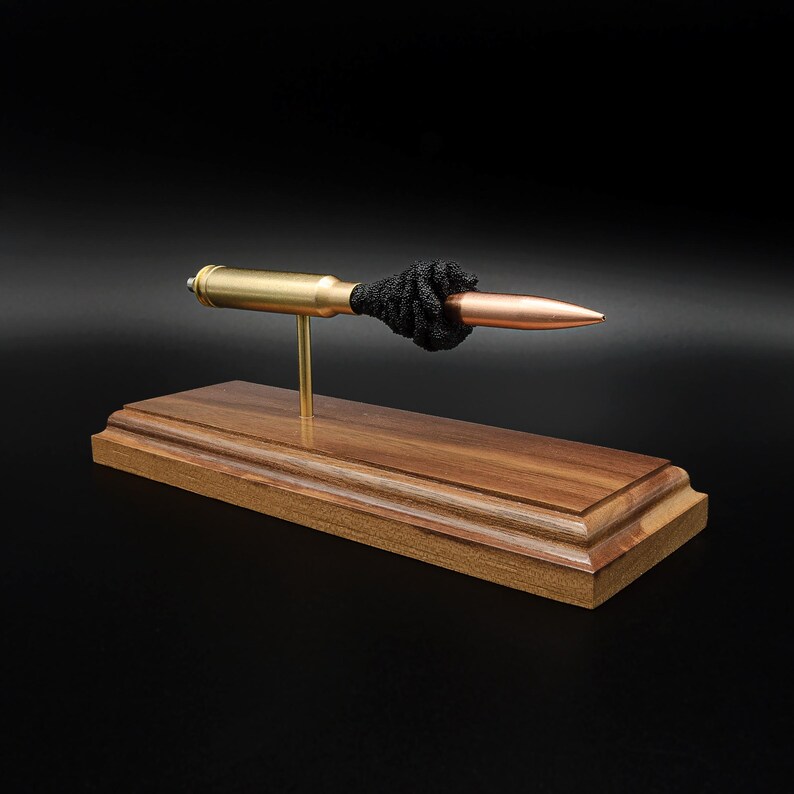Puede incluir: Un expositor decorativo que presenta un casquillo de bala horizontal de lat&oacute;n y cobre montado sobre una fina varilla de metal dorado. La bala est&aacute; acentuada con un detalle de nudo negro texturizado y descansa sobre una base rectangular de madera marr&oacute;n pulida.