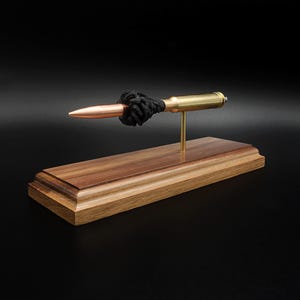 Puede incluir: Un bol&iacute;grafo con forma de bala que presenta una punta de color cobre y un cuerpo de color lat&oacute;n, acentuado con un cord&oacute;n negro anudado. El bol&iacute;grafo est&aacute; montado horizontalmente sobre un poste de lat&oacute;n en una base de madera pulida.