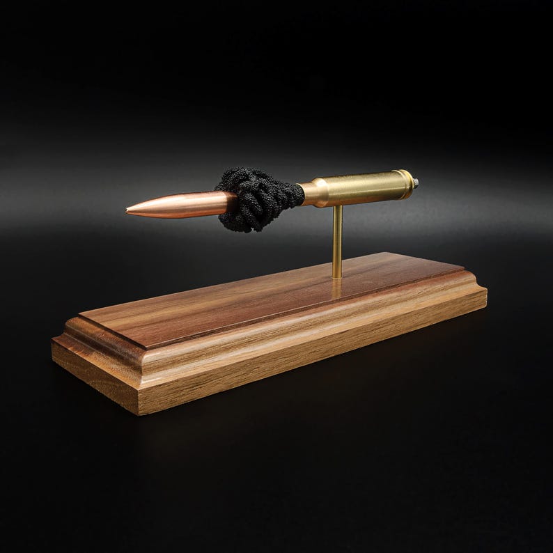 Puede incluir: Un bol&iacute;grafo con forma de bala que presenta una punta de color cobre y un cuerpo de color lat&oacute;n, acentuado con un cord&oacute;n negro anudado. El bol&iacute;grafo est&aacute; montado horizontalmente sobre un poste de lat&oacute;n en una base de madera pulida.