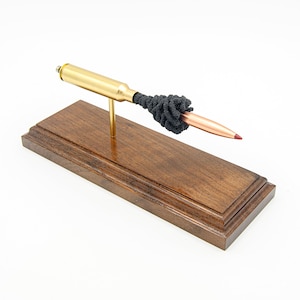 May include: A brass bullet casing mounted on a wooden stand with a black cleaning brush attached to the end. The bullet is displayed on a dark brown wooden base.