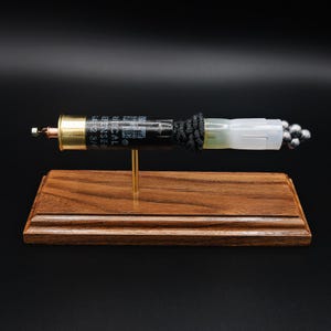 May include: A decorative display featuring a black shotgun shell casing with brass base, connected to a translucent white plastic cylinder filled with small lead-colored spheres, wrapped in black cord, mounted on a rectangular wooden base.