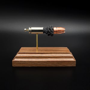 May include: A metallic bullet casing with a copper-colored projectile tip, featuring a cluster of black textured material between the casing and the tip, mounted on a thin gold-toned rod attached to a rectangular wooden base with beveled edges.