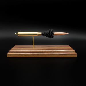 Puede incluir: Un expositor decorativo que presenta un casquillo de bala de lat&oacute;n y una punta de proyectil de color cobre conectados por un cord&oacute;n negro texturizado, montado horizontalmente sobre un poste de metal dorado fijado a una base de madera rectangular.
