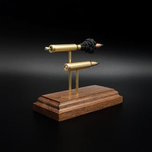 May include: A decorative display featuring two brass bullet casings mounted on thin metal rods atop a rectangular wooden base. The top casing is adorned with a cluster of small black beads and a green-tipped pencil point, while the lower casing is plain.