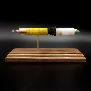 May include: A horizontal pen crafted from a yellow shotgun shell casing with a brass base, a black textured grip, and a white plastic barrel section. The pen is mounted on a thin gold-colored metal rod attached to a rectangular wooden base.