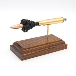 May include: A spent .50 caliber bullet casing mounted on a wooden base. The bullet is filled with black powder and is displayed as a decorative piece.