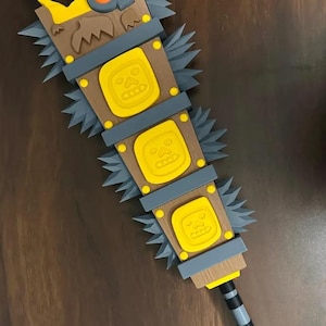 May include: A detailed, close-up view of a fantasy weapon, a mace. The mace features a wooden handle and head with yellow and gray accents. The head has three yellow squares with a face design. The weapon has gray spikes.