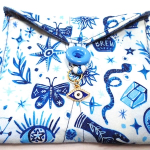 May include: A white fabric pouch featuring a blue ink-style print of butterflies, snakes, stars, crystals, and eyes. The envelope-style flap is secured with a light blue button and a gold-toned metal evil eye charm hanging from a small loop.