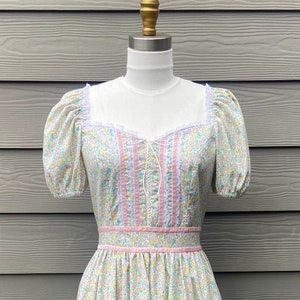 Puede incluir: Un vestido vintage de estampado floral con ribete de encaje rosa y blanco. El vestido tiene un corpi&ntilde;o ajustado y una falda completa con un volante escalonado en el dobladillo.