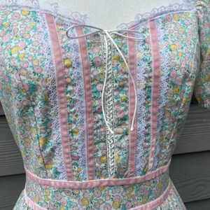 Puede incluir: Un vestido blanco con estampado floral rosa y blanco. El vestido tiene un ribete de encaje y un lazo de cinta rosa en el escote. El vestido tiene un corpi&ntilde;o ajustado y una falda completa.