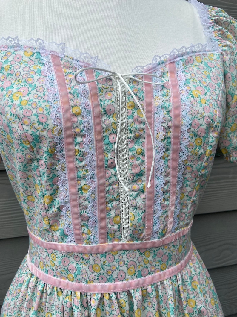 Puede incluir: Un vestido blanco con estampado floral rosa y blanco. El vestido tiene un ribete de encaje y un lazo de cinta rosa en el escote. El vestido tiene un corpi&ntilde;o ajustado y una falda completa.