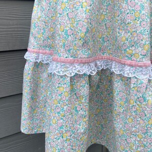 Puede incluir: Una falda blanca con estampado floral en tonos rosa, amarillo y verde. La falda tiene una cinta rosa y un ribete de encaje blanco.