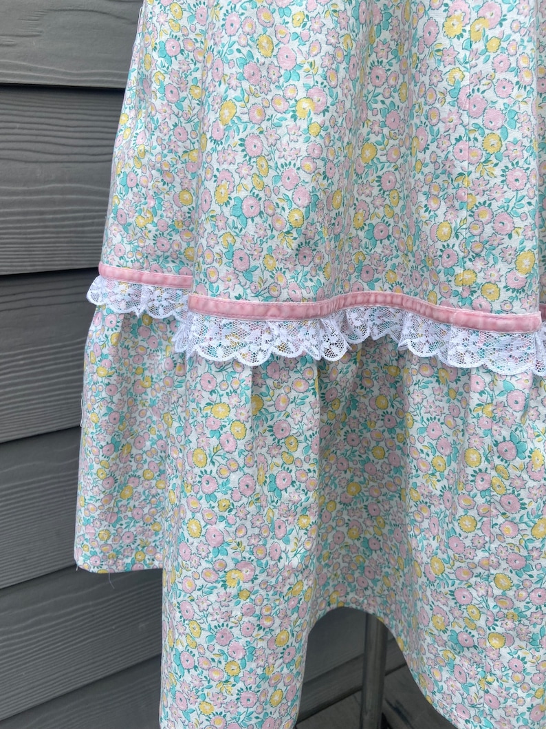 Puede incluir: Una falda blanca con estampado floral en tonos rosa, amarillo y verde. La falda tiene una cinta rosa y un ribete de encaje blanco.