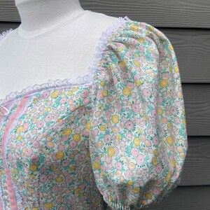 Puede incluir: Un vestido blanco con estampado floral en tonos rosa, amarillo y verde. El vestido tiene un escote cuadrado y mangas abullonadas cortas con ribete de encaje. El vestido tiene un ribete de cinta rosa alrededor del corpi&ntilde;o y un ribete de encaje blanco alrededor del escote.