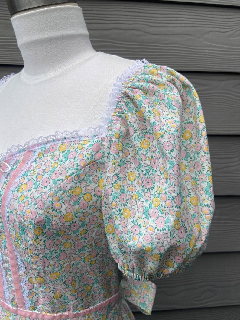 Puede incluir: Un vestido blanco con estampado floral en tonos rosa, amarillo y verde. El vestido tiene un escote cuadrado y mangas abullonadas cortas con ribete de encaje. El vestido tiene un ribete de cinta rosa alrededor del corpi&ntilde;o y un ribete de encaje blanco alrededor del escote.