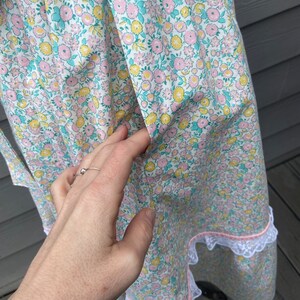 Puede incluir: Un delantal blanco con estampado floral rosa y amarillo y ribete de encaje blanco. El delantal tiene un bolsillo en la parte delantera.