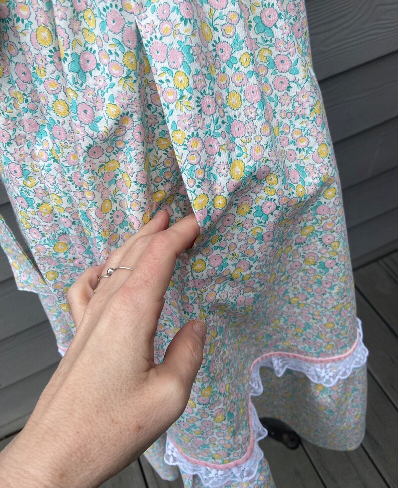 Puede incluir: Un delantal blanco con estampado floral rosa y amarillo y ribete de encaje blanco. El delantal tiene un bolsillo en la parte delantera.