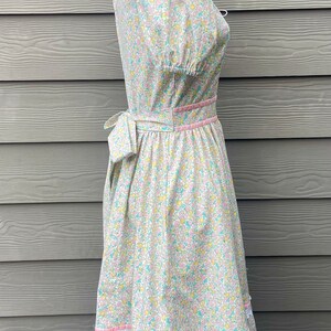 Puede incluir: Un vestido blanco con estampado floral en tonos rosa, amarillo y azul. El vestido tiene un corpi&ntilde;o ajustado y una falda completa con un ribete de encaje. El vestido est&aacute; atado a la cintura con una cinta rosa.