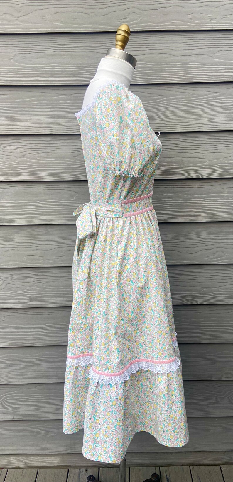 Puede incluir: Un vestido blanco con estampado floral en tonos rosa, amarillo y azul. El vestido tiene un corpi&ntilde;o ajustado y una falda completa con un ribete de encaje. El vestido est&aacute; atado a la cintura con una cinta rosa.