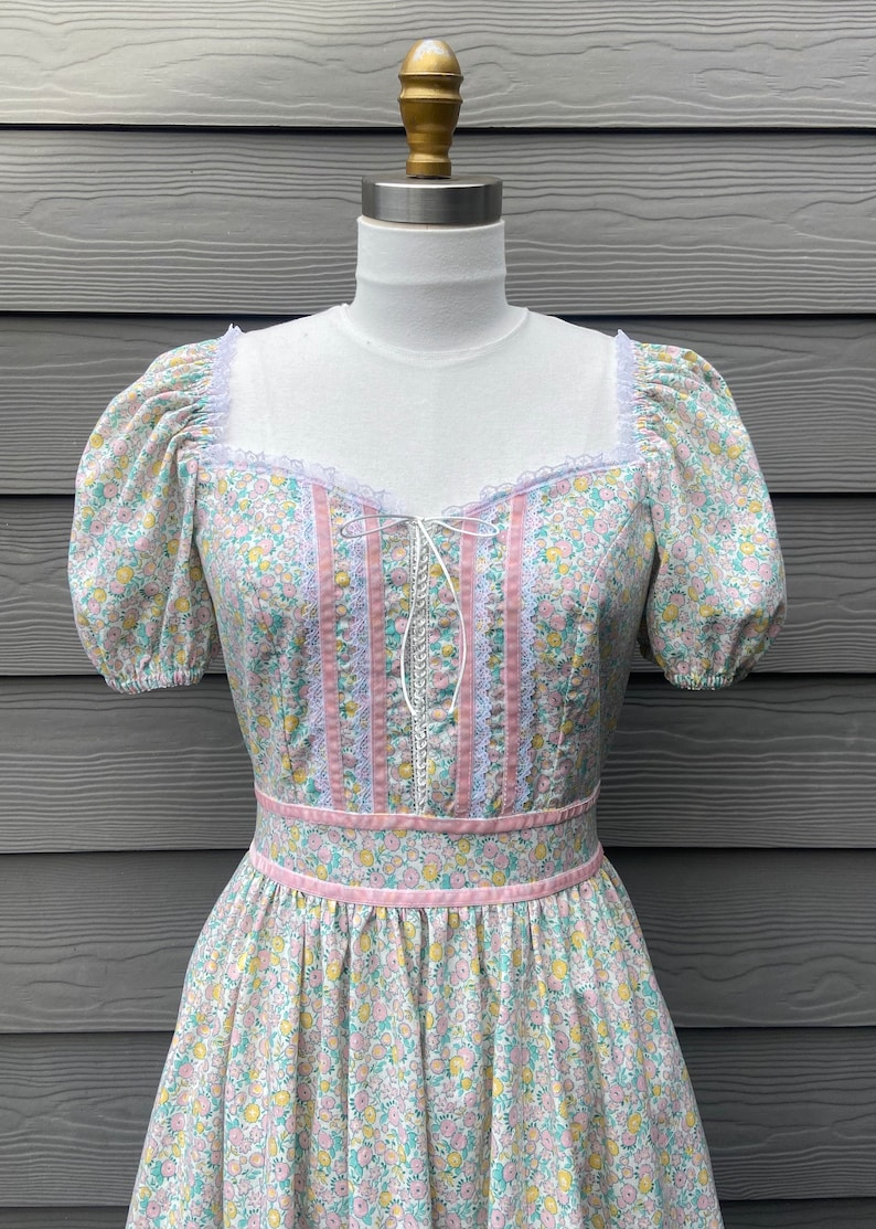 Puede incluir: Un vestido vintage floral con ribete de encaje rosa y blanco, mangas abullonadas y un corpi&ntilde;o ajustado. El vestido tiene un fondo verde claro con un peque&ntilde;o estampado floral.