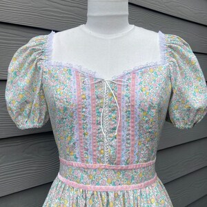 Puede incluir: Un vestido vintage floral con ribete rosa y blanco. El vestido tiene un escote de coraz&oacute;n y mangas abullonadas cortas. La tela es un algod&oacute;n ligero con un peque&ntilde;o estampado floral.