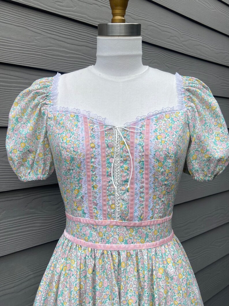 Puede incluir: Un vestido vintage floral con ribete rosa y blanco. El vestido tiene un escote de coraz&oacute;n y mangas abullonadas cortas. La tela es un algod&oacute;n ligero con un peque&ntilde;o estampado floral.
