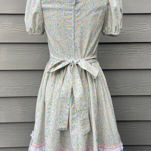 Puede incluir: Un vestido floral azul claro y blanco con escote cuadrado y mangas abullonadas cortas. El vestido tiene un ribete de encaje blanco alrededor del escote y una cinta rosa atada a la cintura. La falda tiene tres capas de ribete de encaje blanco.