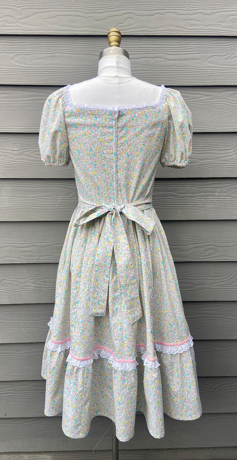 Puede incluir: Un vestido floral azul claro y blanco con escote cuadrado y mangas abullonadas cortas. El vestido tiene un ribete de encaje blanco alrededor del escote y una cinta rosa atada a la cintura. La falda tiene tres capas de ribete de encaje blanco.