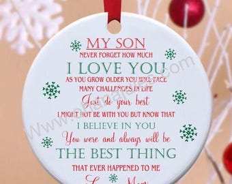 May include: A round white ceramic ornament hanging from a red ribbon, featuring green and red text that reads: MY SON NEVER FORGET HOW MUCH I LOVE YOU AS YOU GROW OLDER YOU WILL FACE MANY CHALLENGES IN LIFE Just do your best I MIGHT NOT BE WITH YOU BUT KNOW THAT I BELIEVE IN YOU You were and always will be THE BEST THING THAT EVER HAPPENED TO ME Love, Mom. Small green snowflakes decorate the text.